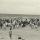 A black-and-white photograph of a crowd of people, most of them standing, on a beach.